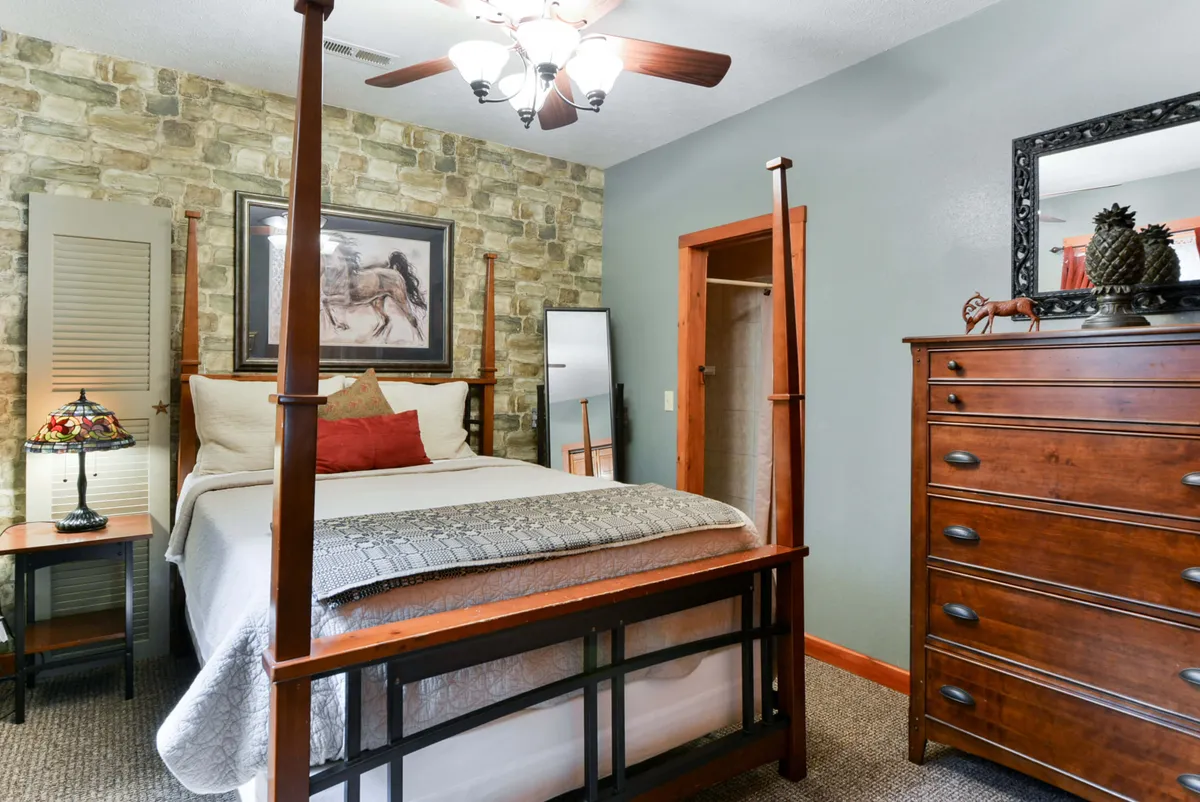 A cozy bedroom featuring a classic four-poster queen bed with elegant posts, a stone accent wall adding rustic charm, a warmly lit table lamp, and a chest of drawers. The bedding looks plush with luxurious linens, and the overall atmosphere is one of comfort and inviting serenity.