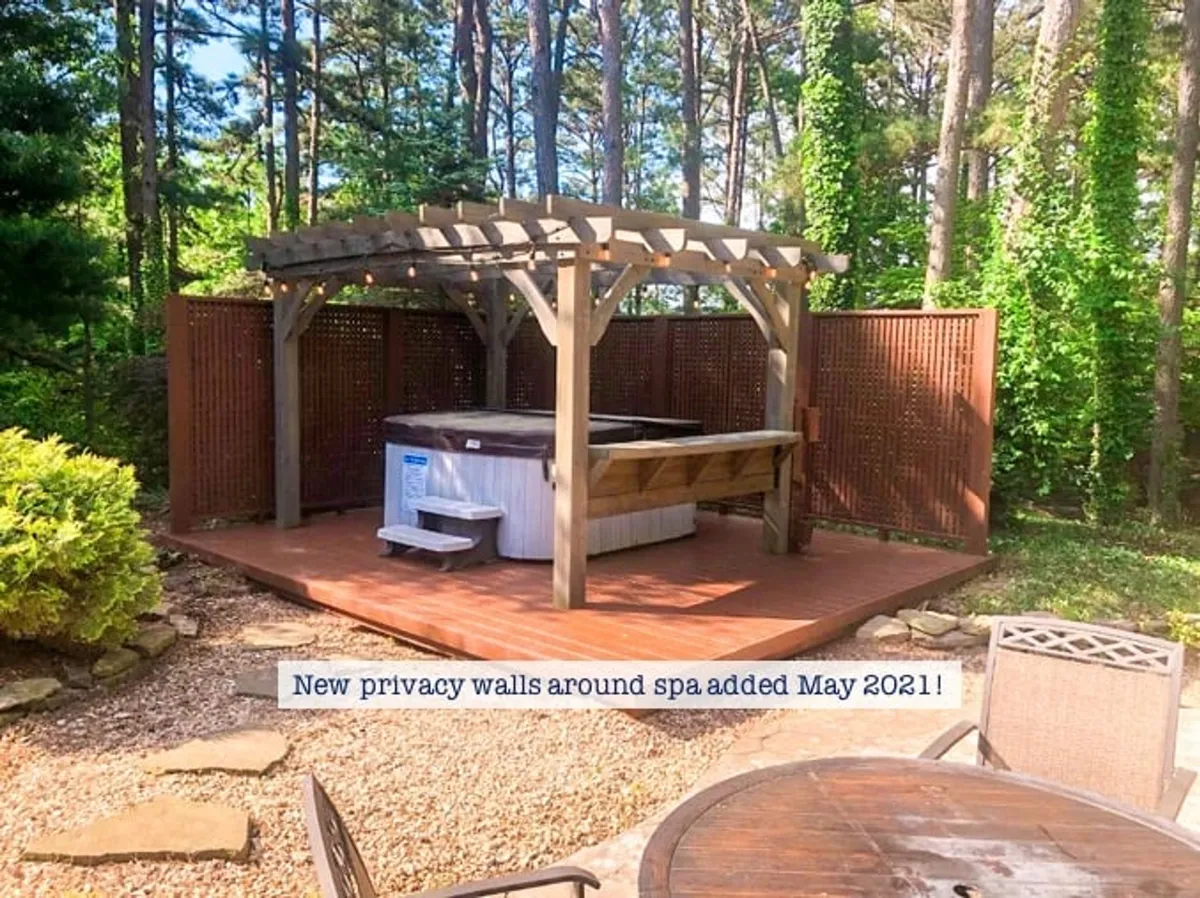 A charming exterior featuring a hot tub under a pergola, flanked by privacy walls and surrounded by trees. The wooden deck invites relaxation and stargazing, perfect for quiet evenings. The setting is intimate yet open to the natural environment, offering both comfort and seclusion.
