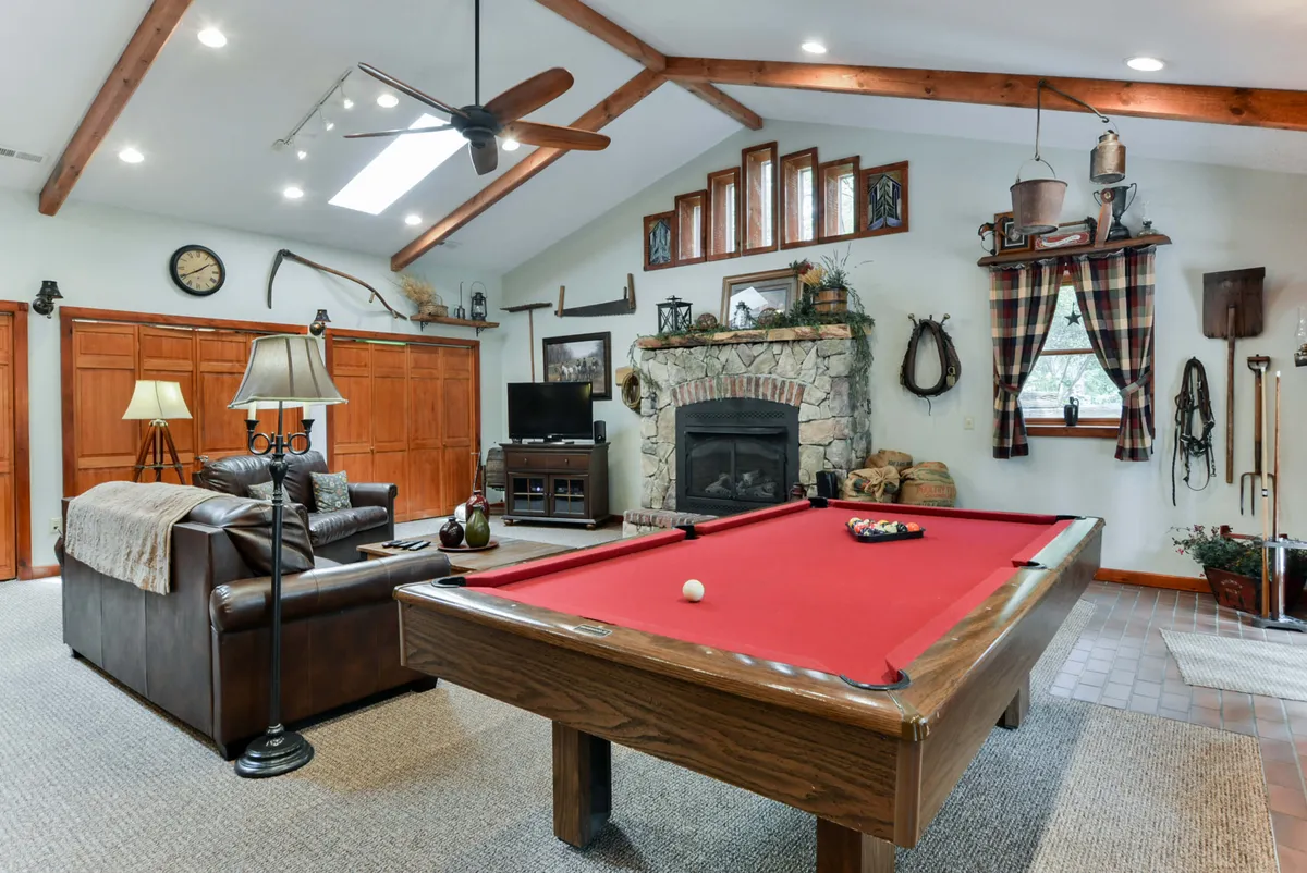 The image features a stylish interior space with a stone fireplace, wooden beams, a pool table, and comfortable seating. The arrangement is ideal for both active play and cozy relaxation, emphasizing elements like family gatherings and game nights.