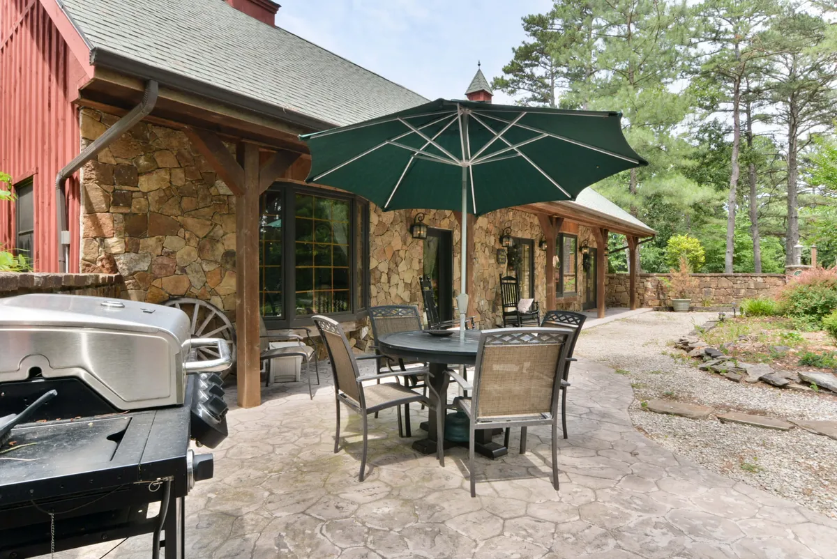 An inviting exterior scene with a BBQ grill, shaded table with umbrella, stone wall, and surrounding greenery. The setup suggests a perfect spot for barbecuing, outdoor dining, and group celebrations in a relaxed, natural environment.