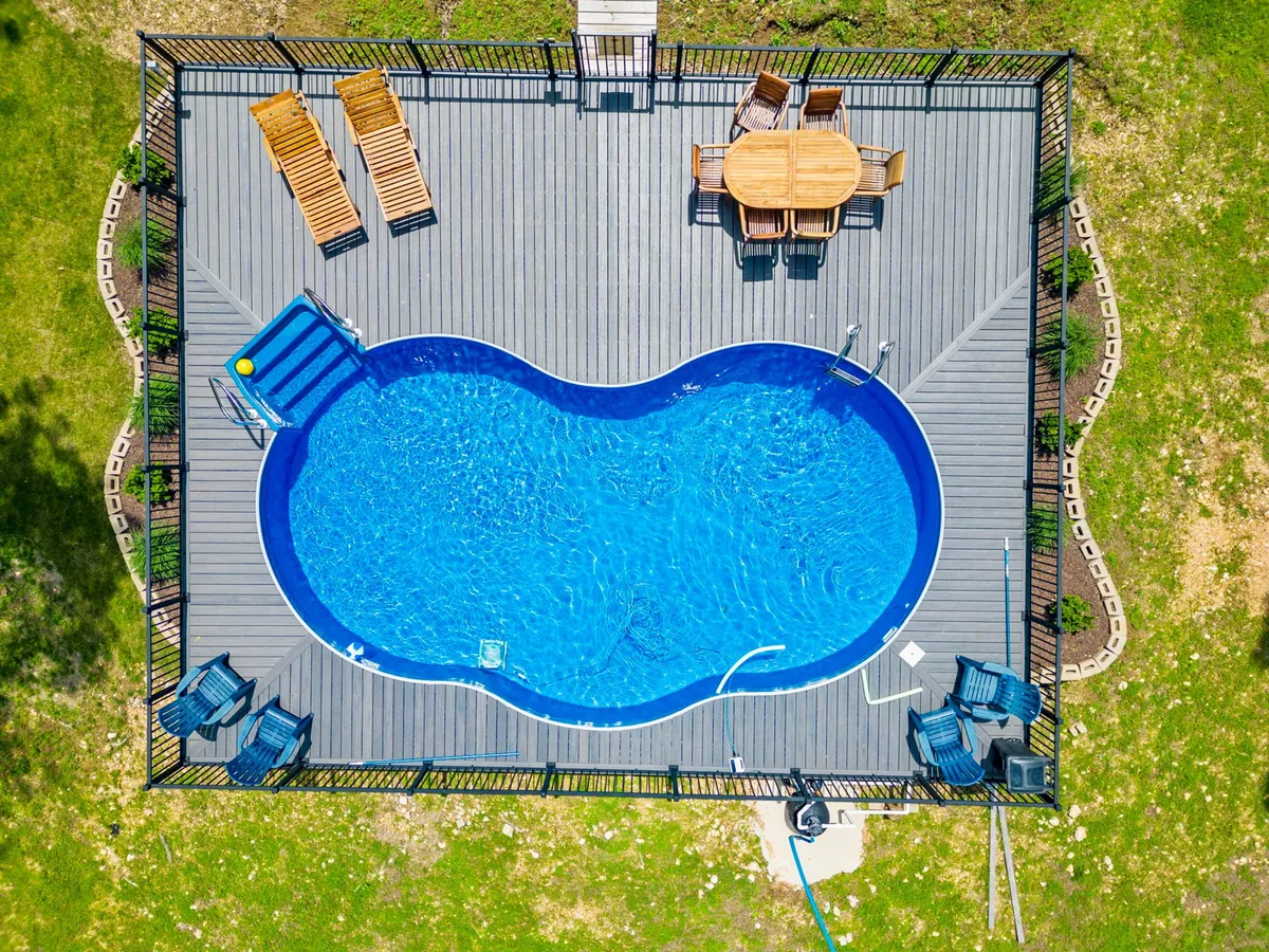 A vibrant exterior shot featuring a sparkling swimming pool surrounded by a deck with outdoor seating and a dining table. The inviting setup is perfect for poolside gatherings, sunbathing, and al fresco meals, all set against lush greenery.