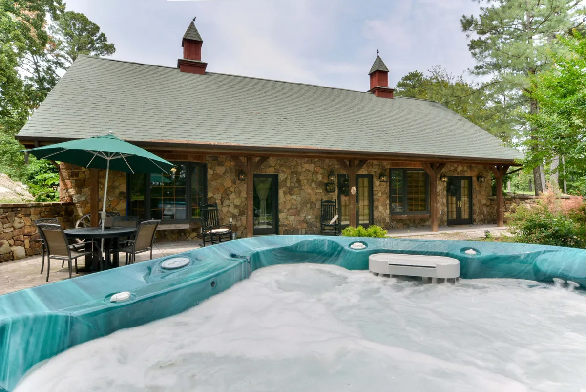 This image features a hot tub set beside an outdoor dining set, with the stone exterior of the house and an umbrella providing shade. The setting is inviting, with space for socializing, dining, and relaxing in the hot tub—enhanced by tranquil surroundings and natural views.