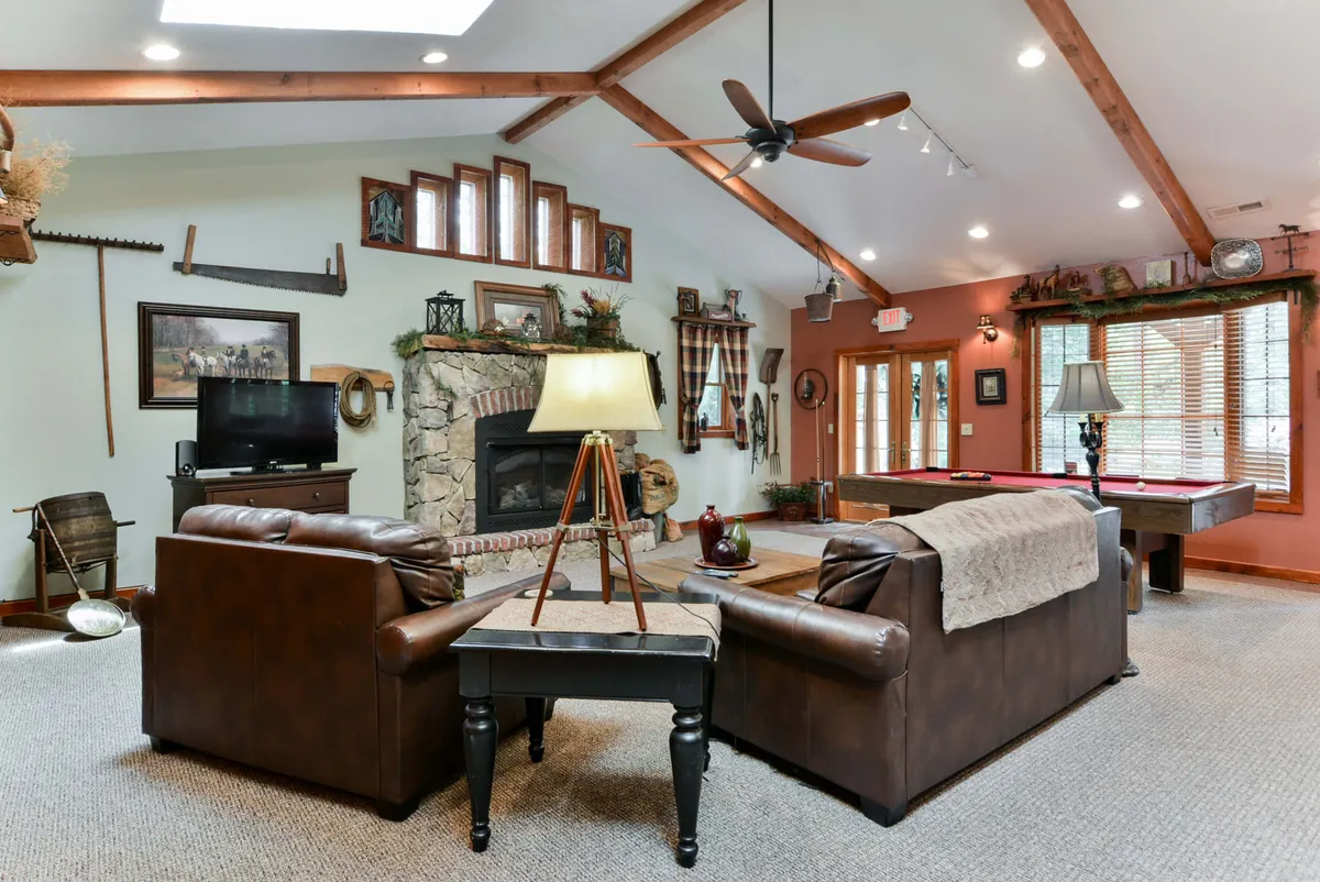 This image showcases a comfortable living room with a prominent fireplace, a pool table, a cozy sofa set, and large windows with blinds that allow natural light to filter in. Decorative wall art adds to the inviting atmosphere, and the space is clearly designed for relaxation, play, and socializing.