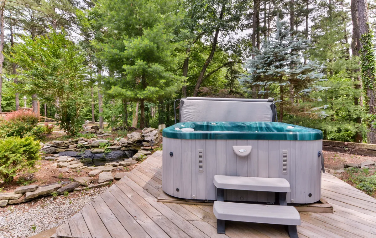 A serene scene showing a hot tub set on wooden decking, surrounded by lush greenery, natural rock formations, and a small pond nearby. The atmosphere is peaceful, offering both relaxation and a close connection to nature.