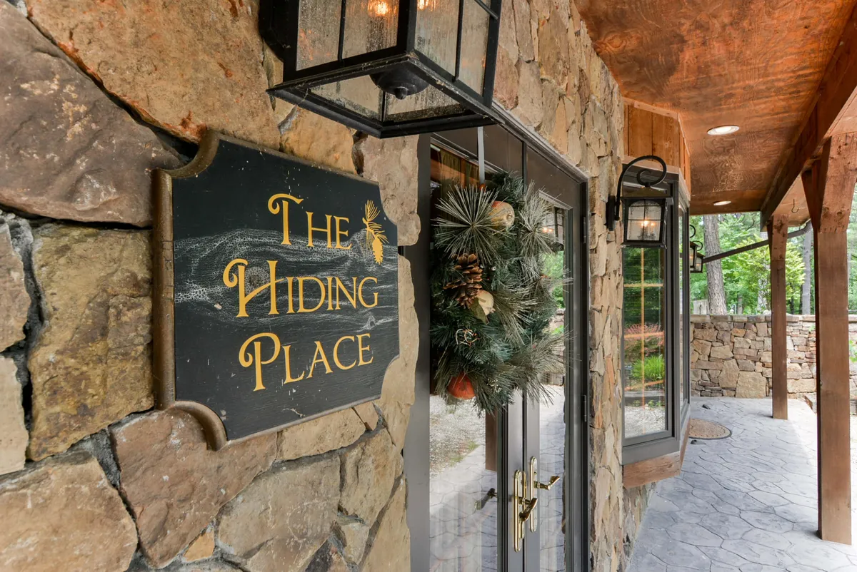 The exterior shot centers on ‘The Hiding Place’ sign with decorative lanterns and a welcoming wreath on the front door, set against a backdrop of trees and outdoor seating. The inviting entry hints at eco-conscious hospitality and a tranquil arrival experience.