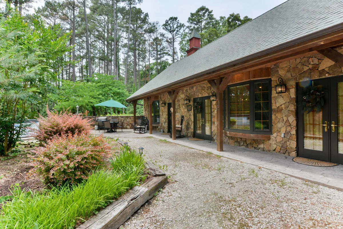 The image features a welcoming stone exterior with wooden beams, lush green landscaping, and a spacious outdoor seating area. The scene feels harmonious and serene, illustrating a comfortable patio where guests can relax with pets and enjoy nature.
