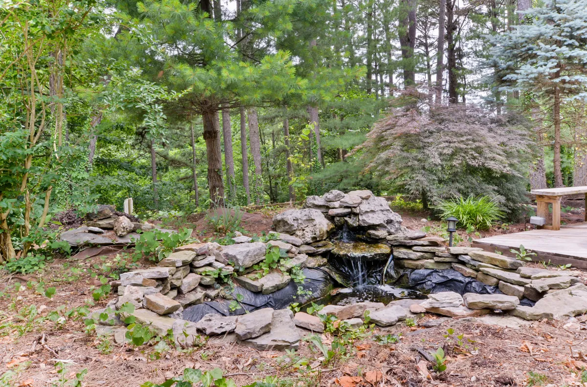 This exterior scene features a stone waterfall nestled among lush greenery, with a wooden bench positioned nearby. The area invites guests to relax by the soothing water, enjoy nature walks, or meditate in the peaceful garden environment.