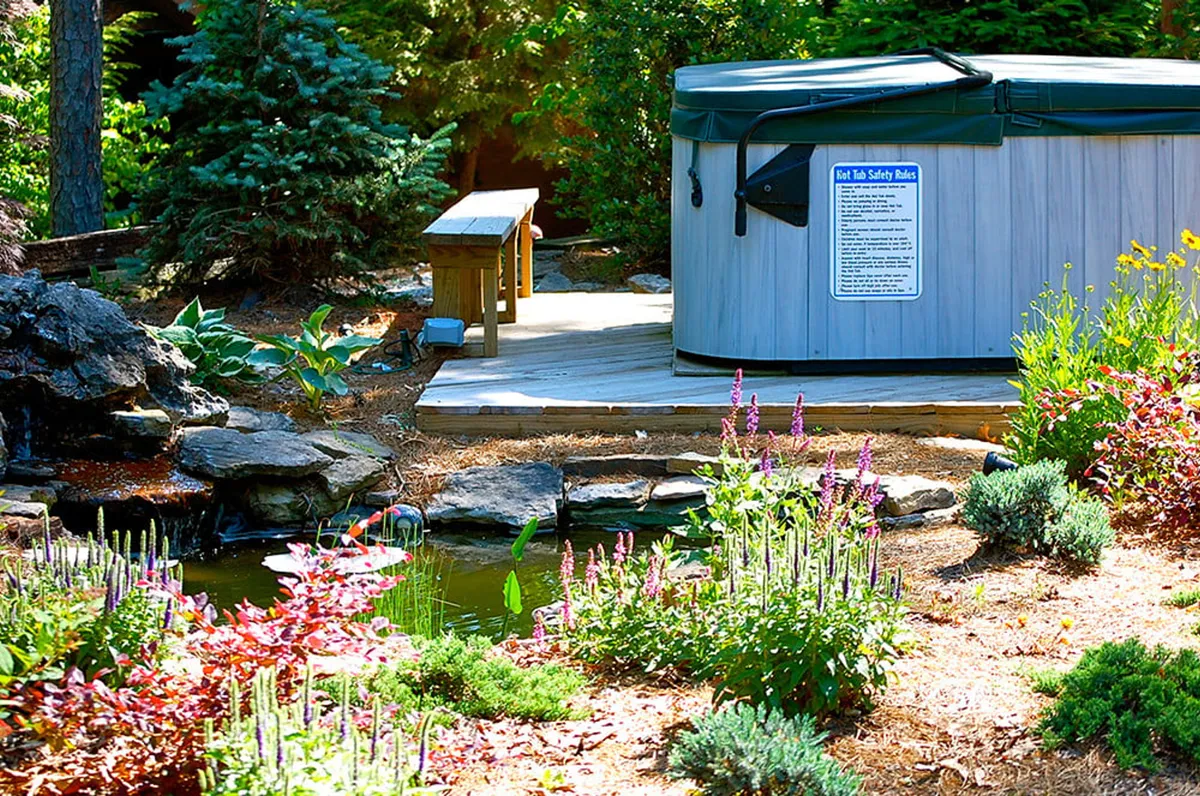 This exterior scene features a hot tub nestled beside a wooden bench, with a rock formation and pond surrounded by flowering plants. The natural landscape evokes privacy and tranquility, ideal for couples seeking a romantic, restorative escape. The scene is softly lit, suggesting an inviting retreat for relaxation and intimate moments.