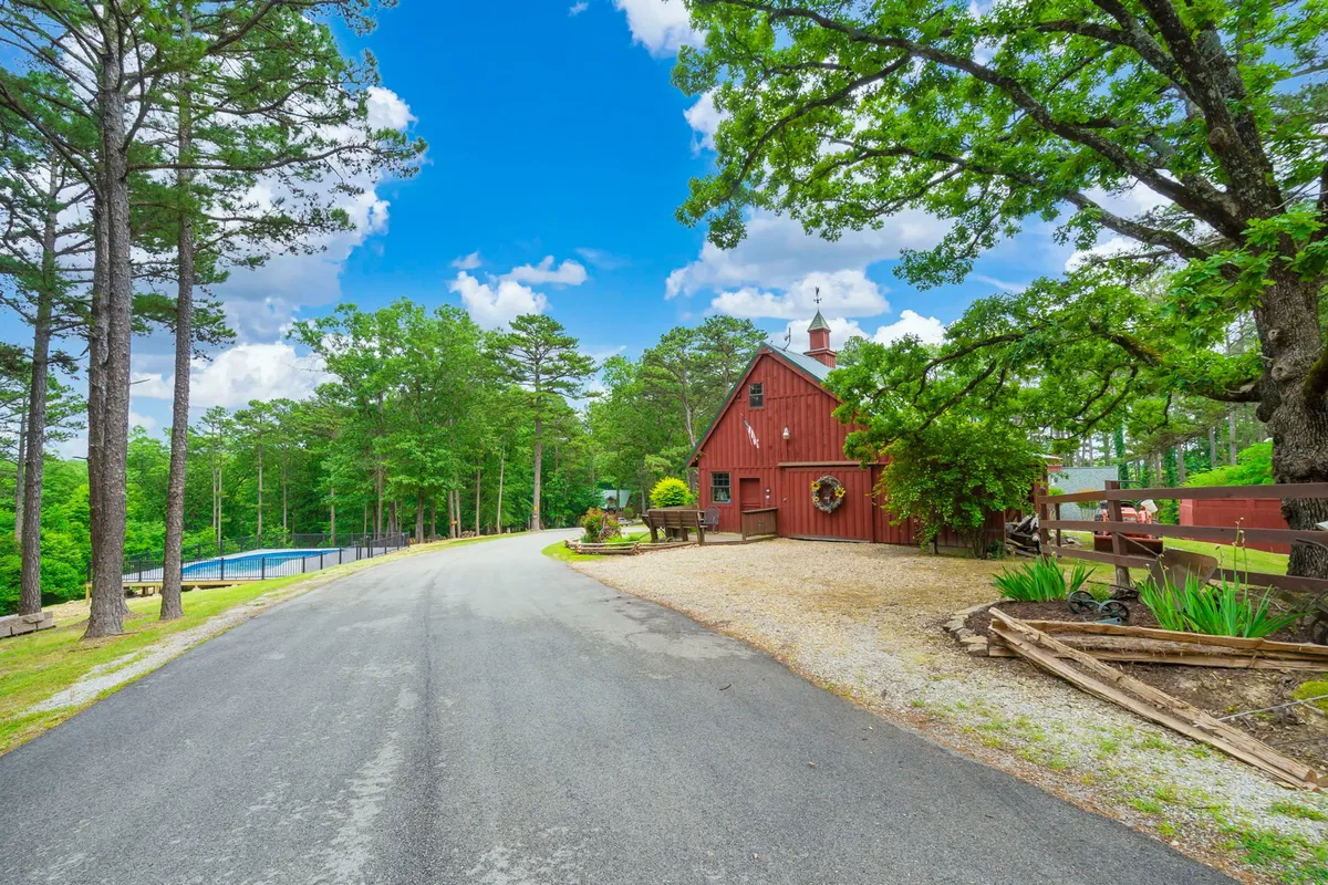 An exterior shot showcasing the red barn-style house with a swimming pool in front, backed by trees and rolling hills. The relaxed scene includes a gravel driveway and expansive nature, offering a sense of removed, peaceful seclusion. The image captures sunset lighting, heightening the romantic atmosphere.